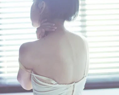 A woman in a light slip with a fallen strap looks away, lit by soft window blinds.