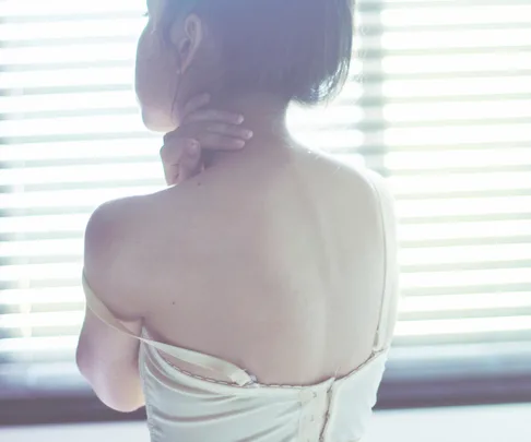 A woman in a light slip with a fallen strap looks away, lit by soft window blinds.