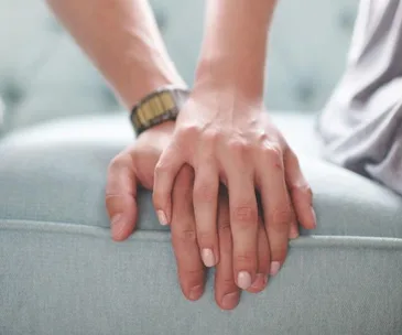 A man wearing a wristwatch holds a woman's hand gently on a light blue sofa.