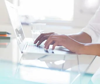 Person typing on a laptop keyboard on a glass desk with a glass of water in the background.