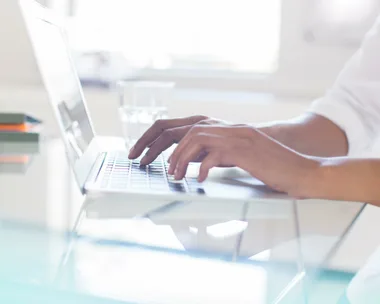 Person typing on a laptop keyboard on a glass desk with a glass of water in the background.