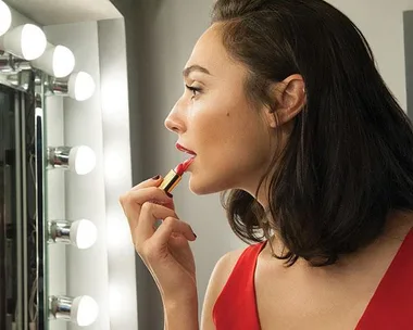 Gal Gadot applying red lipstick in front of a mirror with lit vanity bulbs, wearing a red dress.