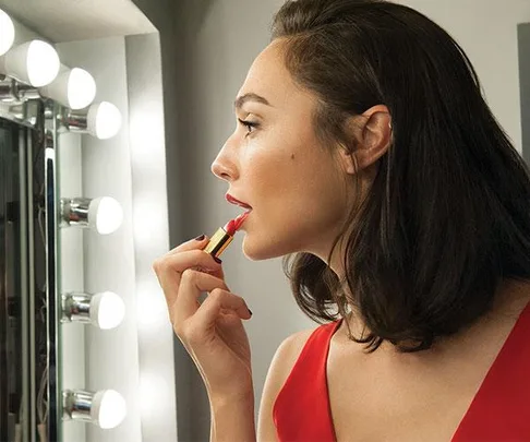 Gal Gadot applying red lipstick in front of a mirror with lit vanity bulbs, wearing a red dress.