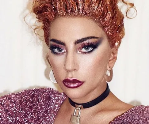 Lady Gaga with styled red hair, bold eye makeup, dark lipstick, wearing a sparkly pink top and a choker with a padlock.