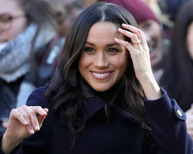 A woman with long dark hair smiling, wearing a dark coat, and touching her hair, with a prominent ring on her left hand.