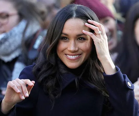 A woman with long dark hair smiling, wearing a dark coat, and touching her hair, with a prominent ring on her left hand.