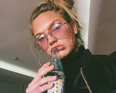 Woman with glasses and hair bun, wearing black jacket, holding bottle, indoors.