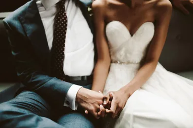 A bride and groom are sitting and holding hands, with the bride in a strapless white dress and the groom in a suit.