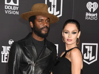 A couple posing on the red carpet at a Justice League event, man in leather jacket and hat, woman in black dress.