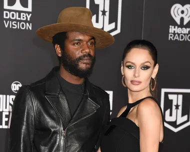 A couple posing on the red carpet at a Justice League event, man in leather jacket and hat, woman in black dress.