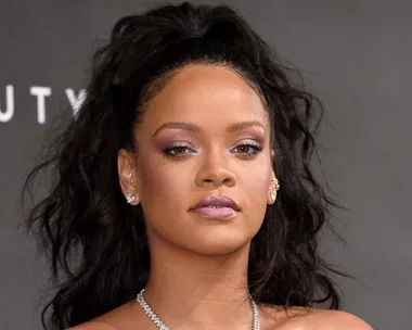 A person with dark long hair, wearing shiny makeup and diamond earrings, against a grey background.