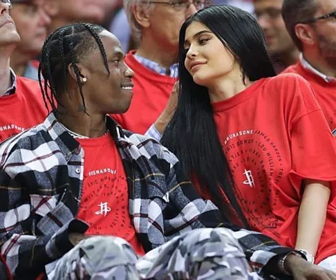 "Kylie Jenner and Travis Scott wearing matching red t-shirts, seated closely and smiling at each other at an event."