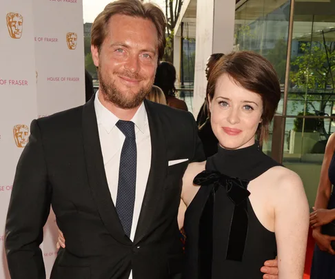 A man in a black suit and tie with short blond hair and beard smiling next to a woman with short brown hair in a black dress.