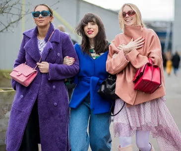 Three women in colorful outerwear, including a purple coat, blue jacket, and peach coat, walking outdoors with bright handbags.