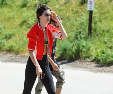 Woman in red jacket and sunglasses hiking on a sunny path with greenery in the background.