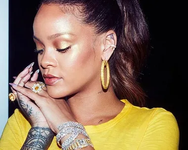 Rihanna wearing a yellow top, gold highlighter, and hoop earrings, showcasing Fenty Beauty products.