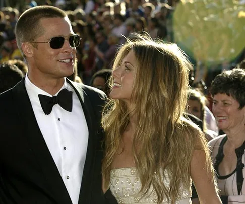 A couple smiling at each other on the red carpet, with the man in a tuxedo and the woman in a white strapless dress, surrounded by people.