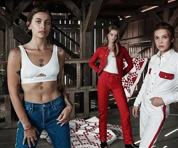 Women wearing Calvin Klein clothing posing in a rustic barn setting with red-and-white quilted blankets in the background.