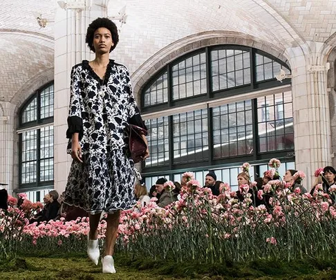 A model walks in a floral black and white dress on a runway adorned with pink flowers, set inside a grand building with tall windows.