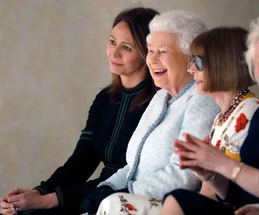 Queen Elizabeth II smiles while attending London Fashion Week, sitting next to two women.