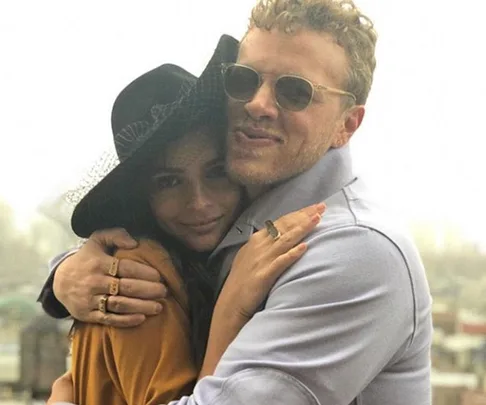 A couple embracing, the woman in a black hat and the man in sunglasses, with a hazy cityscape background.