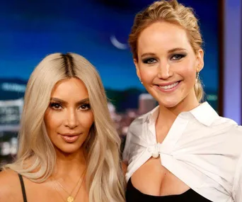 Kim Kardashian and Jennifer Lawrence smiling together on a set with a nighttime city background.