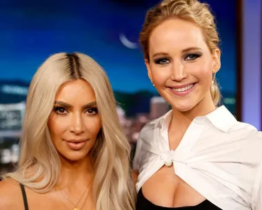 Kim Kardashian and Jennifer Lawrence smiling together on a set with a nighttime city background.