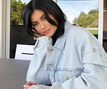 A woman with long dark hair wearing a light denim jacket, resting her head on her hand, with a blurred background.