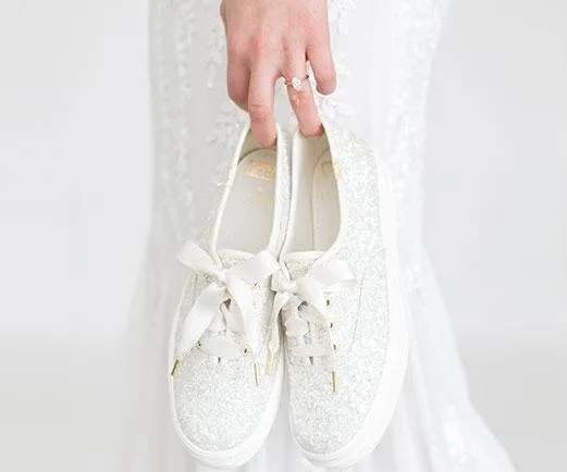 Bride holding pair of white glittery wedding sneakers with satin laces by Keds x Kate Spade New York, wearing a lace wedding dress.