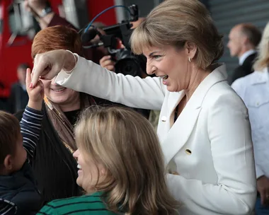 Michaelia Cash smiling and pointing while interacting with a child, surrounded by other people and a camera crew.