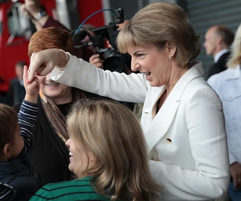 Michaelia Cash smiling and pointing while interacting with a child, surrounded by other people and a camera crew.