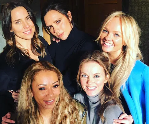 The Spice Girls are smiling and posing together for a group photo indoors.