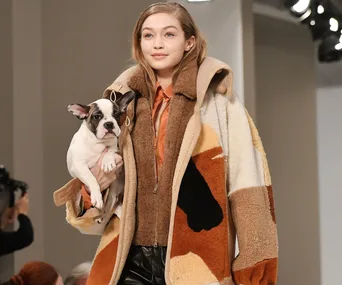 Model holding a French Bulldog, wearing a stylish patchwork coat, smiling on the runway at Tod's Fall 2018 show.