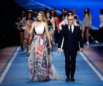 Gigi Hadid and Tommy Hilfiger walk down the runway for Tommy Hilfiger show; models in vibrant outfits follow behind.