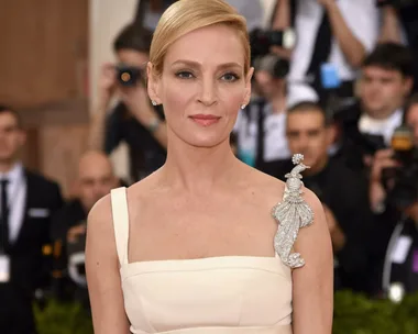 A woman in a beige dress with a diamond-studded brooch on her shoulder, attending an event with photographers in the background.