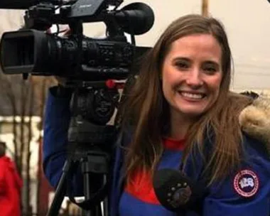 Woman smiling while holding a professional video camera, wearing a blue jacket with a furry hood in an outdoor setting.