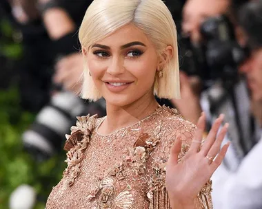 Kylie Jenner on the red carpet, wearing a floral-embroidered dress and waving with cameras in the background.