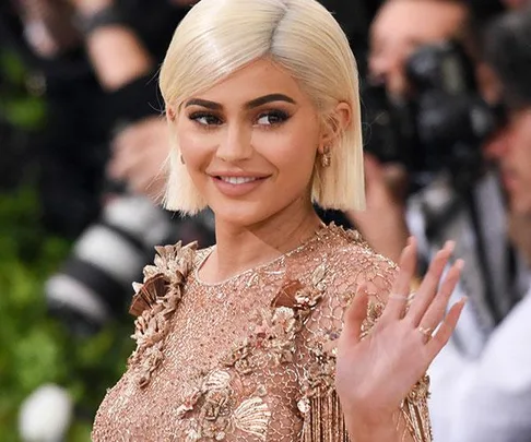 Kylie Jenner on the red carpet, wearing a floral-embroidered dress and waving with cameras in the background.