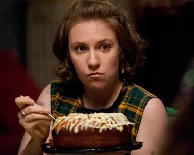 A woman with short, brown hair sits at a table, looking serious, while holding a plate with a frosted cake and a fork.