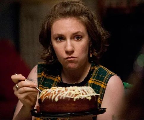 A woman with short, brown hair sits at a table, looking serious, while holding a plate with a frosted cake and a fork.