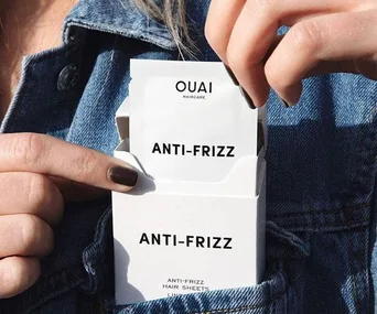 Person pulling out a Ouai Anti-Frizz Hair Sheet from a white box in their denim jacket pocket.