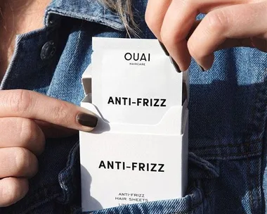 Person pulling out a Ouai Anti-Frizz Hair Sheet from a white box in their denim jacket pocket.