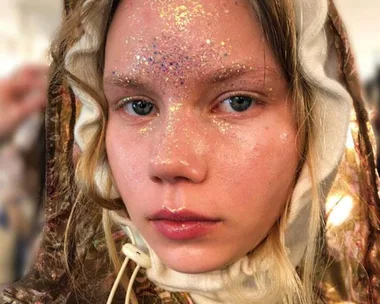 Close-up of a model's face with glitter makeup on forehead and cheeks, wearing a detailed hooded garment.