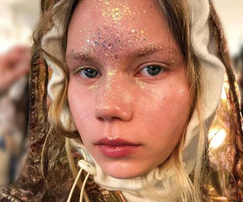 Close-up of a model's face with glitter makeup on forehead and cheeks, wearing a detailed hooded garment.