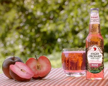 Bottle and glass of Angry Orchard Rosé hard cider with sliced apples on a checkered cloth with blurred green background.