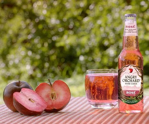 Bottle and glass of Angry Orchard Rosé hard cider with sliced apples on a checkered cloth with blurred green background.