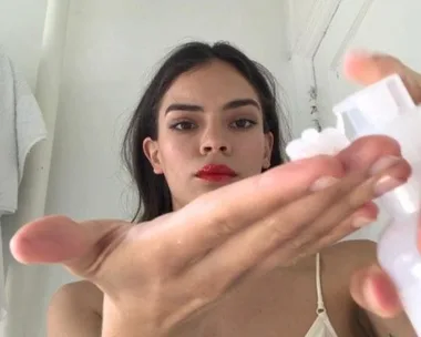 Woman with red lipstick applying skincare product to her palm from a pump bottle in a bathroom.