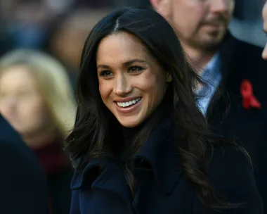 Meghan Markle smiling, wearing a dark coat, with a blurred crowd in the background.