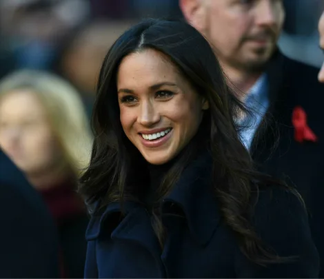 Meghan Markle smiling, wearing a dark coat, with a blurred crowd in the background.