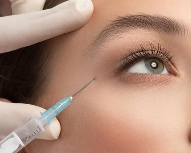 A close-up of a woman receiving an anti-aging injection near her eye, with gloved hands holding the syringe.